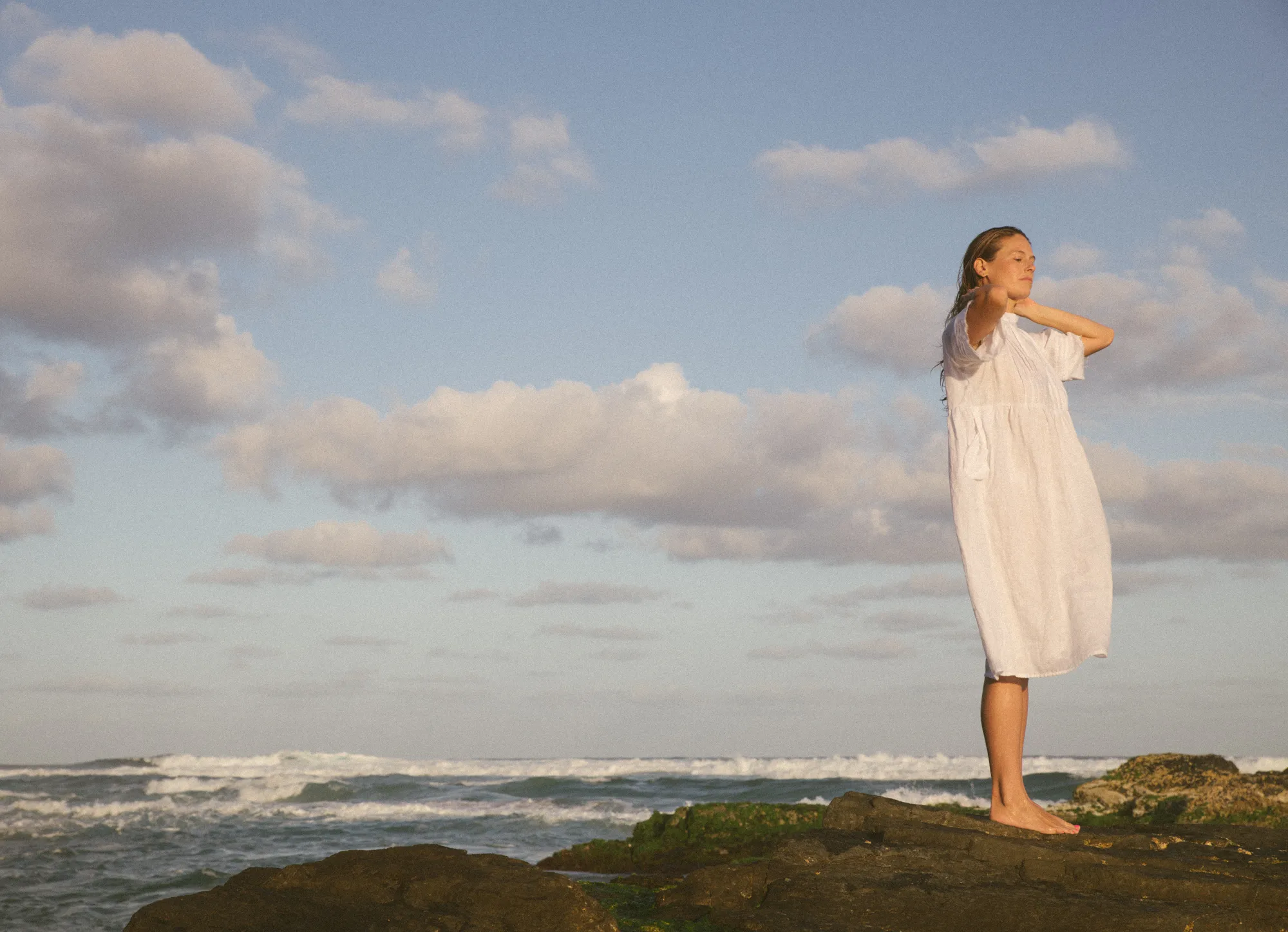 A woman wearing a white dress stands on a rocky ledge. In the background, the ocean waves are crashing.